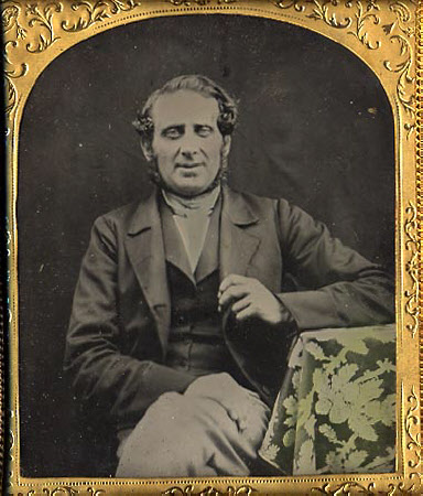 Gentleman seated, his left elbow resting on as table and holding the lapel of his jacket with his ...