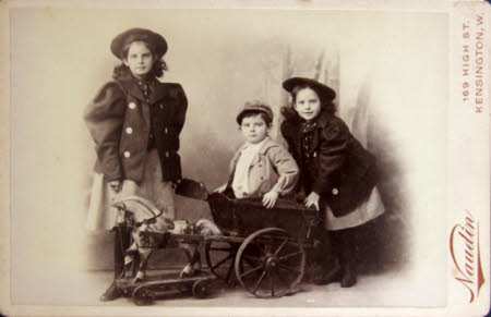 Two girls dressed in jackets with leg of mutton sleeves and a boy kneeling in a toy horse and cart ...