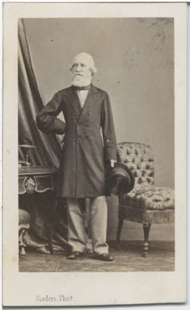 A portrait of an old, bearded gentleman in a shirt and long coat, holding a top hat.