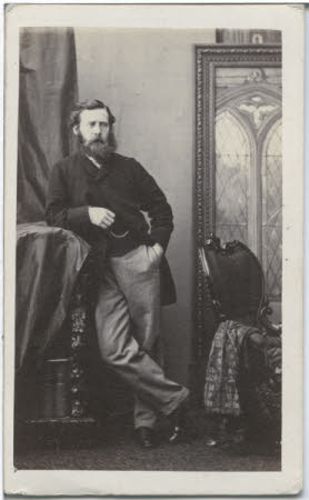 A portrait of a bearded man in formal wear, leant against an ornate bookcase.